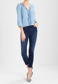 Hellbluse mit rüschenbesetztem Ausschnitt, Dreiviertelärmeln, kombiniert mit dunkelblauen Skinny-Jeans und silbernen High Heels.