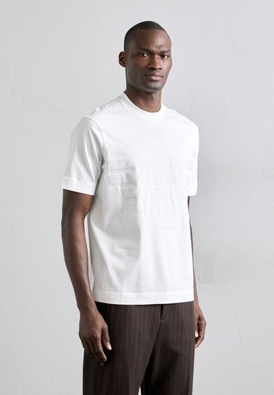 T-shirt in cotone bianco con maniche corte, caratterizzato da un design del logo testurizzato sul davanti. Abbinato a pantaloni marroni a righe verticali.