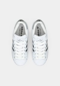 Baskets blanches en cuir avec un embout texturé, intégrant trois rayures noires, des lacets blancs et un logo Adidas sur la languette et les semelles intérieures.