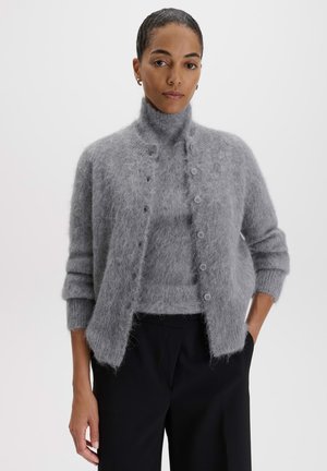 Donna che indossa un cardigan grigio peloso abbottonato sopra un maglione dolcevita abbinato con pantaloni neri, in piedi con una mano in tasca.