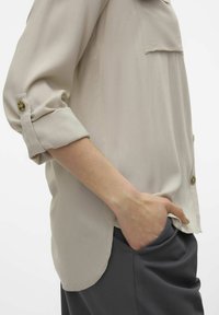 Blusa beige chiaro con maniche arrotolate, tasca frontale e vestibilità rilassata, abbinata a pantaloni grigio scuro. Tessuto liscio, dettagli minimi.