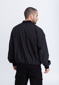 Schwarze Bomberjacke mit gerippten Bündchen und Saum, aus einem glatten, leichten Stoff und lockerem Schnitt. Die Rückansicht zeigt ein schlichtes Design.