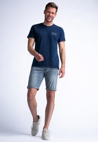 Navyblauwe short-sleeve t-shirt met witte letters, gecombineerd met lichtdenim shorts. Het model draagt lichte sneakers en staat op een lichte achtergrond.