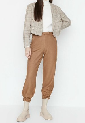 Veste en peluche beige à carreaux, col roulé blanc, pantalon brun fuselé avec poignets élastiques et bottes à talons compensés beiges.