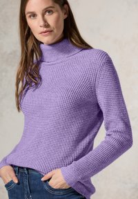 Pull à col roulé en tricot violet avec un motif texturé, manches longues et poignets côtelés. Porté avec un jean en denim bleu, les mains dans les poches.