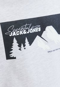T-shirt gris avec un design graphique noir représentant des montagnes et des arbres, accompagné du texte « Essentials JACK & JONES » en blanc.