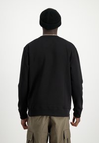 Felpa nera con una vestibilità classica, polsini e orlo a coste, texture liscia, e una piccola patch con logo sul retro, abbinata a pantaloni cargo beige.