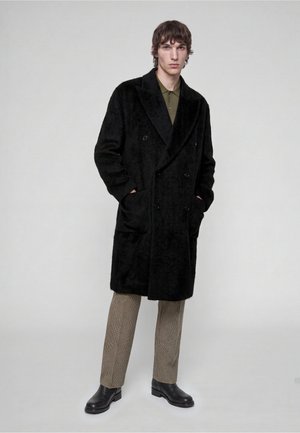 Mann trägt einen schwarzen, doppelreihigen Mantel, olivgrünes Hemd, karierte Hose und schwarze Stiefel, steht mit den Händen in den Taschen.