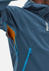 Veste imperméable bleue avec doublure orange. Comprend des poches zippées avec des accents bleus, des poignets ajustables et un col haut. Tissu léger.