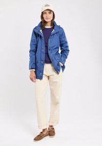 Giacca impermeabile blu con cappuccio, interno bianco, indossata sopra una maglia viola. Abbinata a pantaloni crema e scarpe marroni, con risvolti.