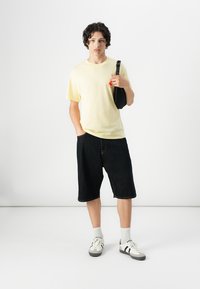 Camiseta de algodón amarilla, pantalones cortos de mezclilla oscura, calcetines blancos y zapatillas en blanco y negro. El modelo sostiene una bolsa negra con una correa estampada.
