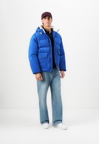 Mand iført blå dunjakke, lyseblå jeans, hvide sneakers og maroon kasket, der står mod en enkel hvid baggrund.