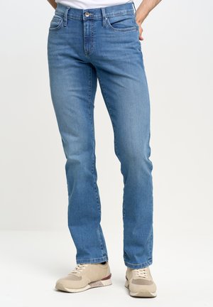 Muž stojící proti jednoduchému světlému pozadí, oblečený v světle modrých džínách s rovným střihem a béžových teniskách.