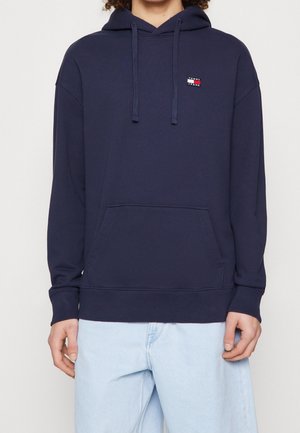 Granatowa bluza z kapturem z przednią kieszenią i logo Tommy Jeans, noszona z jasnoniebieskimi dżinsami na osobie na jednolitym tle.