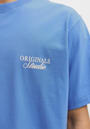 Sininen puuvillapaita, jossa on pyöreä kaula-aukko ja lyhyet hihat. Edessä on valkoista brodeerattua tekstiä: "ORIGINALS Studio."