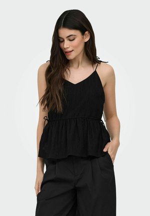 Jeune femme aux longs cheveux foncés portant un haut noir sans manches avec des bretelles à nouer et un pantalon noir, posant avec une main dans la poche.