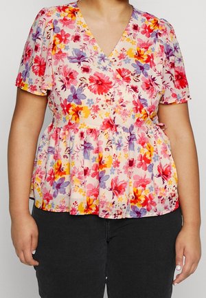 Donna che indossa una camicetta floreale a maniche corte con scollo a V decorata con fiori rossi, viola e gialli, abbinata a pantaloni neri.