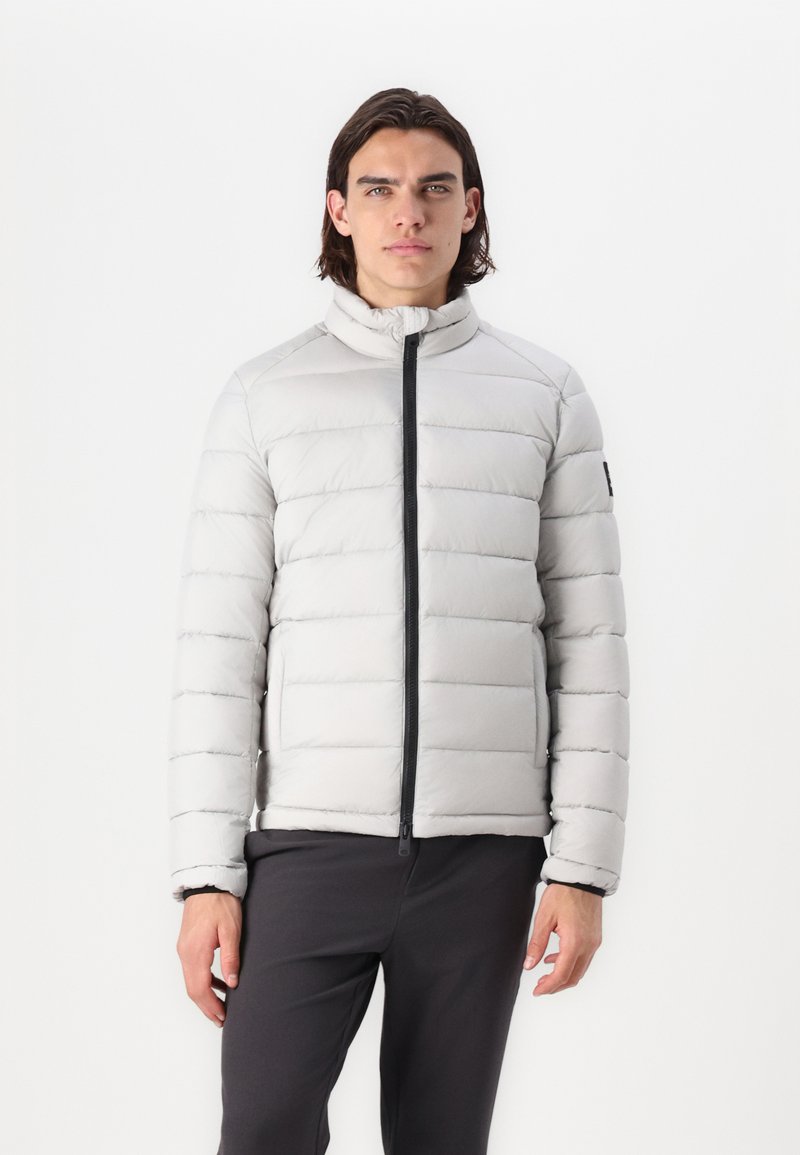 Chaqueta acolchada de color gris claro con un alto cuello, costuras horizontales, cremallera frontal completa y bolsillos laterales. Textura suave y diseño ajustado.