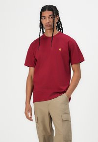 Red cotton t-shirt with short sleeves and a crew neck. Features a small gold logo on the chest. Paired with beige cargo pants.