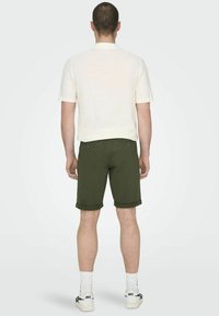 Camisa polo de crema para hombre con mangas cortas, combinada con pantalones cortos enrollados de color verde oscuro, calcetines blancos y zapatillas con acentos negros.