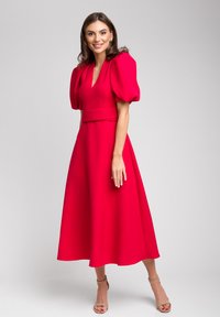 Czerwona sukienka do kolan z bufiastymi rękawami, dekoltem w kształcie litery V i dopasowaną talią. Gładka tekstura i zwiewny krój. Noszona z cielistymi szpilkami.