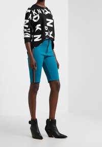 Persoon die een zwart-witte DKNY-trui draagt, teal korte broek tot aan de knie met oranje streep, en zwarte enkelhoge laarzen met hak, staand in een witte kamer.