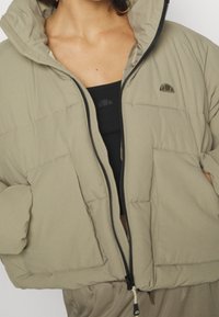 Cropped puffer jacket in light khaki, featuring a zip front, large pockets, and a high collar. Smooth, padded texture with logo detail.