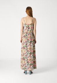 Femme aux longs cheveux roux portant une robe maxi fleurie sans manches et des sandales bleues à talons, debout de dos sur un fond blanc uni.