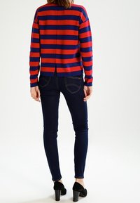 Czerwono-granatowy sweter w paski o luźnym kroju, z rozporkiem z tyłu, zestawiony z ciemnymi wąskimi dżinsami i czarnymi butami na obcasie.