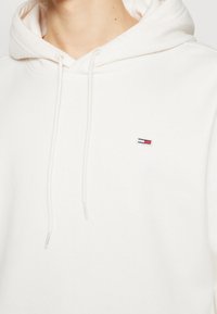 Primer plano de una persona que lleva una sudadera con capucha blanca con un pequeño logotipo rectangular rojo, blanco y azul en el pecho.