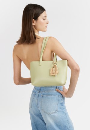 Donna con capelli castani fino alle spalle che porta una borsa verde pallido con un portachiavi intrecciato, indossa un top senza spalline e jeans blu a vita alta.