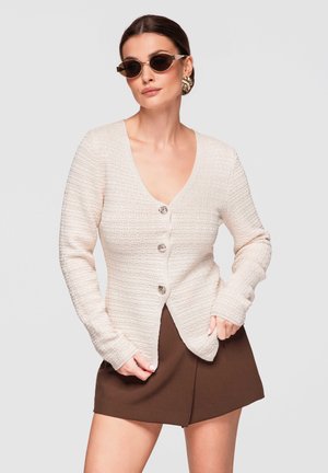 Kobieta w beżowym, teksturowanym kardiganie na guziki, brązowej spódnicy, okrągłych okularach przeciwsłonecznych i dużych złotych kolczykach, pozująca na tle jednolitego jasnego tła.