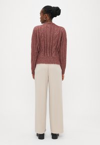 Πλεκτό πουλόβερ με σχέδιο καλωδίων σε βουργουνδί απόχρωση με ριμπ τελείωμα, φορεμένο πάνω από εκρού παντελόνι με φαρδιά πόδια. Η υφή του νήματος προσθέτει βάθος, αναδεικνύοντας το σχέδιο.