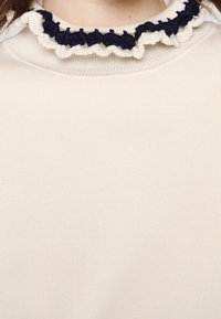 Rippensweater in Creme mit einem schwarzen und beigen, voluminösen Häkelkragen. Der Stoff wirkt weich, hat eine glatte Textur und ist figurbetont geschnitten.