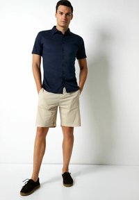 Marineblaues kurzärmliges Hemd mit Knöpfen, beigefarbene Shorts und dunkle Schnürschuhe. Glattes Material mit taillierter Passform; die Schuhe haben eine geflochtene Sohle.