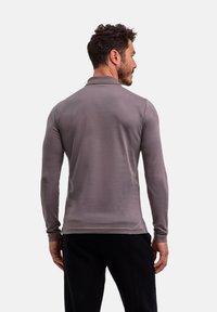 Koszulka polo z długim rękawem w jasnoszarym materiale z kołnierzykiem, z bezszwowym tyłem i minimalnymi przeszyciami. Noszone są czarne spodnie.