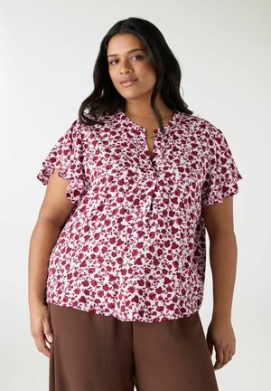 Donna che indossa una camicetta bianca con stampa floreale rossa e maniche a volant, abbinata a pantaloni marroni, in piedi davanti a uno sfondo semplice.