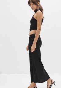 Schwarzes, ärmelloses Crop-Top, kombiniert mit einem langen schwarzen Rock, der eine geraffte Detail an der Taille und einen silbernen Akzentknopf hat.