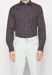 Homme portant une chemise à manches longues à motifs floraux sombres, rentrée dans un jean taille haute bleu clair.