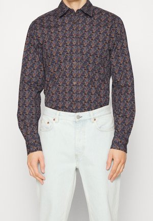Homme portant une chemise à manches longues à motifs floraux sombres, rentrée dans un jean taille haute bleu clair.