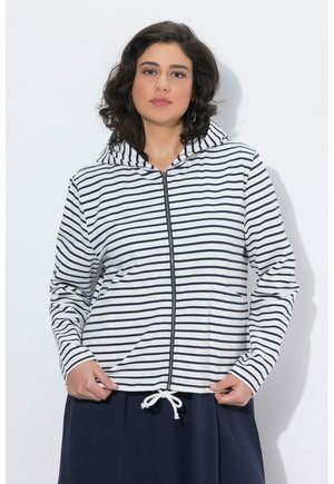 Femme portant un sweat à capuche blanc à fermeture éclair avec des rayures horizontales noires et un cordon de serrage à l'ourlet, assorti à une jupe foncée.