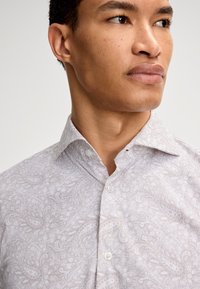 Hellgraues Hemd mit Paisleymuster, aus Baumwollstoff, strukturierter Kragen und weißen Knöpfen. Nahaufnahme des Ausschnitts und Schulterbereichs.