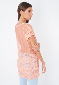 Top de melocotón semitransparente con bordado floral, mangas cortas y espalda descubierta. Textura suave y diseño ligero, combinado con jeans de mezclilla claros.