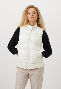 Puffy cream vest with a high collar, zip closure, and green logo detail. Worn over a black long-sleeve top, paired with light beige pants.