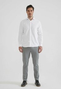 Chemise blanche à manches longues avec un col boutonné, associée à un pantalon tailleur gris clair et des chaussures noires à lacets. Design simple, tissu lisse.