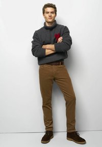 Pull grise avec une broderie en forme de cœur rouge, pantalon en velours côtelé marron et bottes en daim marron. Le modèle pose les bras croisés devant un fond neutre.