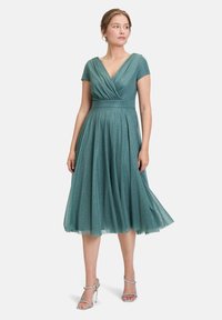Robe en tulle verte avec un décolleté en V et des manches courtes, présentant une taille ajustée et une jupe fluide ornée de subtiles paillettes.