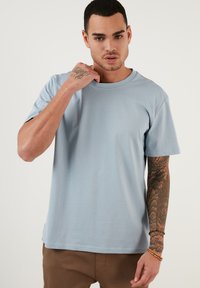 Tricou din bumbac de culoare albastru deschis, cu guler rotund, mâneci scurte și o croială relaxată. Prezintă tatuaje vizibile pe un braț și o textură netedă.