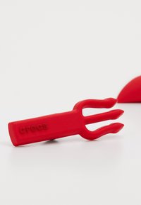 Roter Gummi-Crocs-Anhänger in Form einer dreizackigen Gabel, mit glatter Textur und eingeprägtem Markennamen.