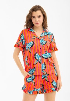 Vrouw draagt een kleurrijke, bijpassende, knoopsluitende top met korte mouwen en korte broek met blauw vogel- en oranje gestreept patroon, staand met één hand in de zak.